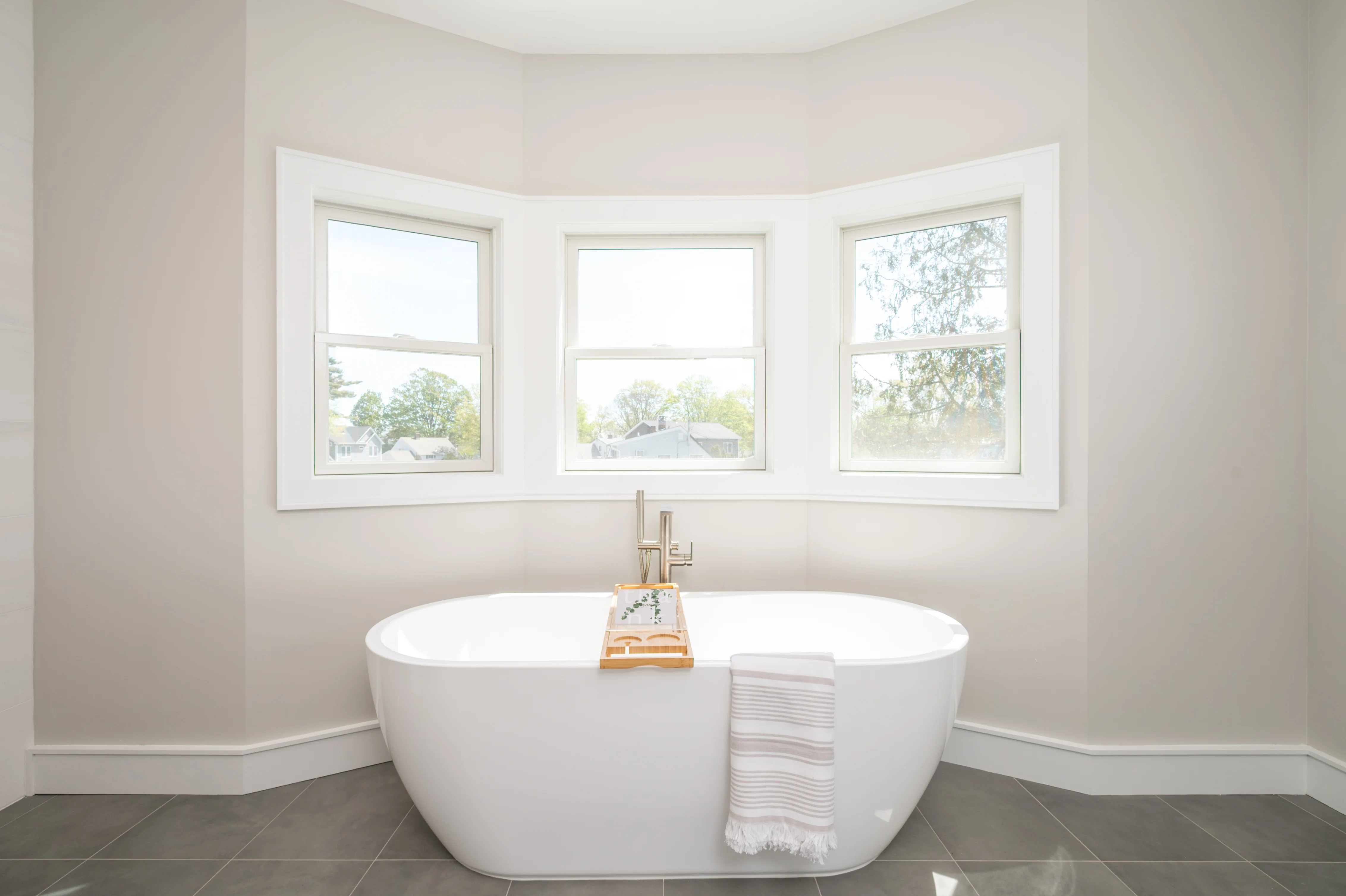 Three white double-hung windows in curved bay above freestanding white soaking tub with tile flooring